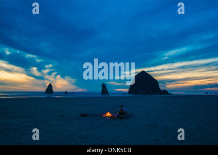 Paar in der Nähe von Lagerfeuer, Haystack Rock & The Needles im Hintergrund, Cannon Beach, Oregon, USA Stockfoto
