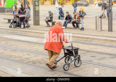 Alte Dame mit Orangen Mantel und Rollator mit der Handbremse, überqueren einer Straße mit Embeded Straßenbahnschienen – Heilbronn Deutschland Stockfoto