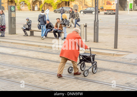 Alte Dame mit Orangen Mantel und Rollator mit der Handbremse, überqueren einer Straße mit Embeded Straßenbahnschienen – Heilbronn Deutschland Stockfoto