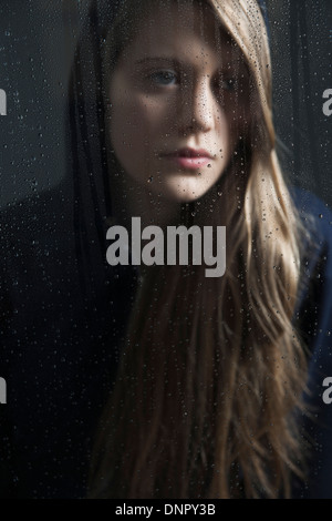 Porträt der jungen Frau hinter Fenster, nass mit Regentropfen, mit hoodie Stockfoto