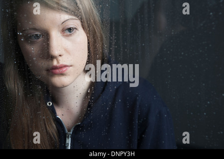 Porträt der jungen Frau hinter Fenster, nass mit Regentropfen, mit hoodie Stockfoto