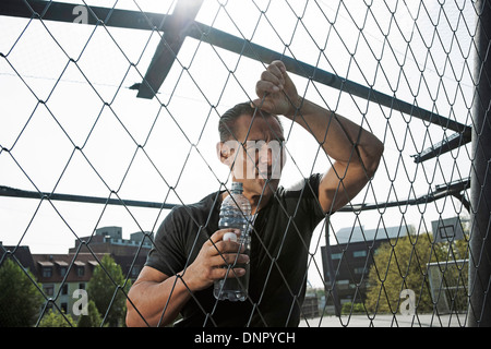 Reifer Mann gelehnt Maschendrahtzaun am Basketballfeld im Freien, halten Sie die Flasche Wasser, Deutschland Stockfoto