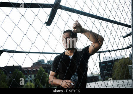 Reifer Mann gelehnt Maschendrahtzaun am Basketballfeld im Freien, halten Sie die Flasche Wasser, Deutschland Stockfoto