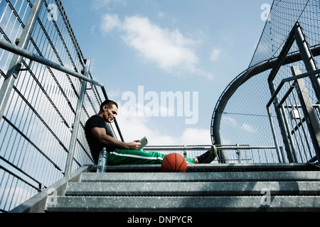 Reifer Mann sitzt an Spitze der Treppe am Basketballfeld im freien Blick auf Tablet-PC, Deutschland Stockfoto