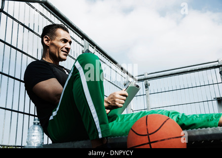 Reifer Mann sitzt an Spitze der Treppe am Basketballfeld im freien Blick auf Tablet-PC, Deutschland Stockfoto