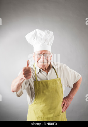 Senior-Mann mit Schürze und Küchenchefs Hut geben Thumbs Up, Studioaufnahme Stockfoto