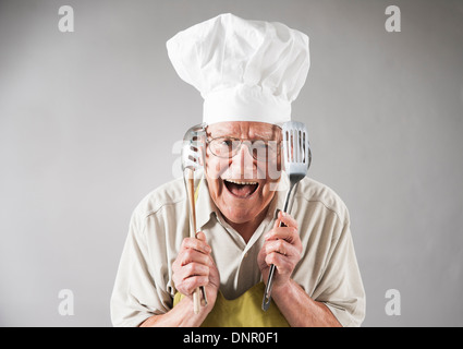 Senior-Mann mit Kochutensilien Hut Schürze und Küchenchefs, Studioaufnahme Stockfoto