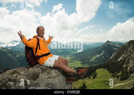 Reifer Mann sitzen auf Klippe mit Luft, erhobenen Armen wandern in Bergen, Tannheimer Tal, Österreich Stockfoto