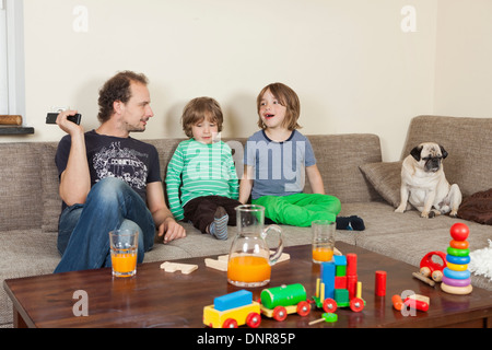 Vater mit Söhnen und Hund auf dem Sofa, im Vordergrund auf einem Tisch Spielzeug und Getränke Stockfoto