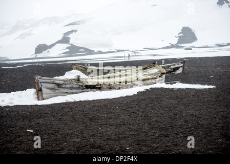Whalers Bay Deception Island Verlassenes Ruderboot Winter | DECEPTION ISLAND, Antarktis — Zwei verfallene Holzflenserruderboote liegen gestrandet auf dem schwarzen Vulkansand von Whalers Bay, Teil der Ruinen der Hektor-Walfangstation, die als Historische Stätte und Denkmal Nr. 71 des Antarktis-Vertrags geschützt ist. Solche Boote brachten Walfänger zwischen dem Ufer und den schwimmenden Fabrikschiffen, die seit der Saison 1906–07 in Port Foster vor Anker lagen, hin und her, ein verschwenderischer Prozess, der bis 1912–13 etwa 3.000 verrottende Kadaver hinterließ, die den Hafen verschmutzten. Stockfoto