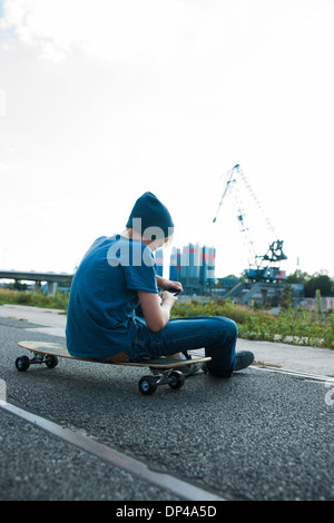 Rückansicht des jungen sitzen auf Skateboard im Freien in einem Industriegebiet und Blick auf Handy, Deutschland Stockfoto