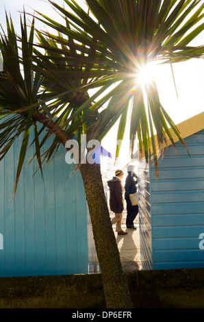 Menschen zu Fuß auf der Promenade zwischen Strandhütten, wenn die Sonne untergeht hinter einer Palme. Stockfoto