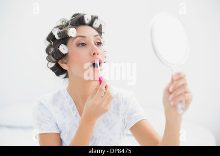 Hübsche Brünette in Lockenwickler halten Handspiegel und Lipgloss auftragen Stockfoto