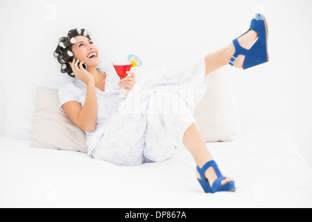 Brünette in Lockenwickler und Keil Schuhe halten einen Cocktail tätigen eines Anrufs auf Bett Stockfoto
