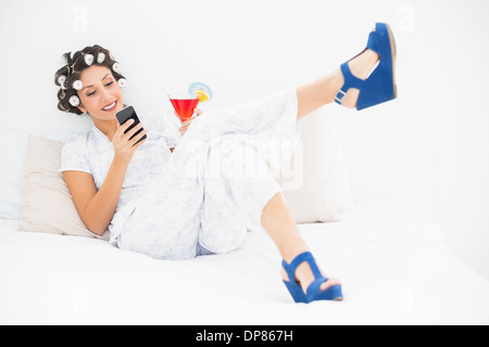 Brünette in Lockenwickler und Keil Schuhe mit einem Cocktail eine SMS auf Bett Stockfoto