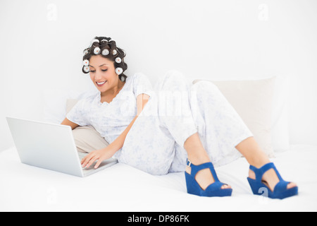 Lächelnde Brünette in Lockenwickler und Keil Schuhe mit ihrem Laptop auf Bett Stockfoto