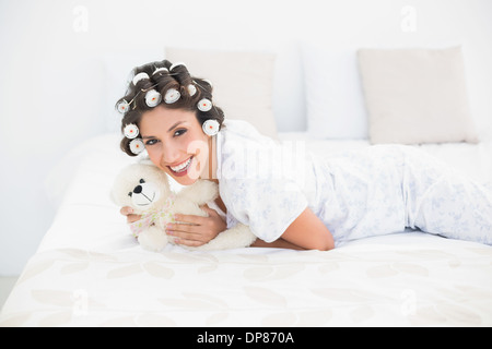 Hübsche Brünette in Lockenwickler den Teddy auf Bett kuscheln Stockfoto