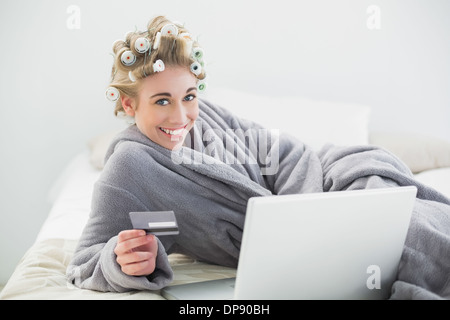 Fröhliche entspannte blonde Frau in Lockenwickler mit ihrem Laptop online kaufen Stockfoto