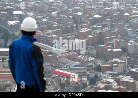 EL ALTO, Bolivien, 9. Januar 2014. Ein Mitarbeiter des Ministeriums für öffentliche Arbeiten beobachtet, wie ein Hubschrauber ein synthetisches Lichtkabel zwischen Pylonen der neuen Seilbahn trägt / Gondel lift derzeit im Bau, die Stadt nach La Paz unten zu verknüpfen. Dies ist der erste Teil des Prozesses, das endgültige Stahlseil zu installieren, das Gondeln tragen wird. Drei separate Routen sollen im Rahmen eines ehrgeizigen Projekts, Verkehrsstaus, mit der ersten Zeile aufgrund zu lindern bis März 2014 abgeschlossen sein. Das rote Gebäude an der Unterseite ist der Bahnhof neben dem Hauptfriedhof. Stockfoto