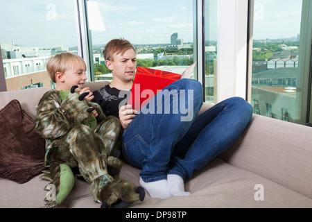 Junge gekleidet in Dinosaurier Kostüm mit Vater Geschichte Buch auf Sofa-Bett zu Hause sitzen Stockfoto