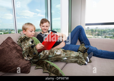 Junge gekleidet in Dinosaurier Kostüm Bilderbuch mit Vater auf Sofa-Bett zu Hause lesen Stockfoto