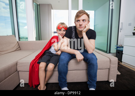 Porträt eines lächelnden jungen gekleidet in Superhelden-Kostüm mit trauriger Vater auf Sofa-Bett zu Hause sitzen Stockfoto