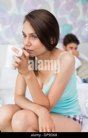 Junge Frau mit Kaffee am Bett mit im Hintergrund liegenden Mann Stockfoto