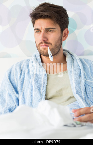 Kranker Mann mit Thermometer im Mund sitzt auf Bett Stockfoto