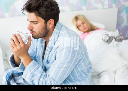 Kranker Mann trinken Kaffee am Bett, während die Frau im Hintergrund zu Hause schlafen Stockfoto