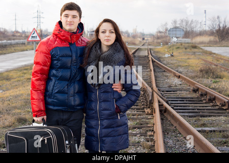 Liebespaar warten für einen Zug stehenden Arm in Arm neben einer ländlichen Eisenbahnlinie mit ihrem Koffer gerade für die seine Ankunft. Stockfoto