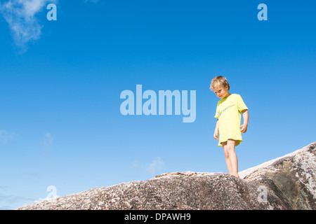 Ein blonder Junge in einem gelben T-shirt auf einen Felsen vor blauem Himmel steht.