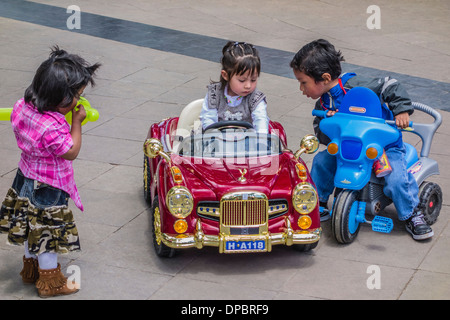 Eine Oberschicht bolivianischen Mädchen sitzt in ihrem sehr extravagant Spielzeugauto während zwei Arme, indigene Kinder an der Plaza 25 de Mayo, Sucre aussehen auf. Stockfoto