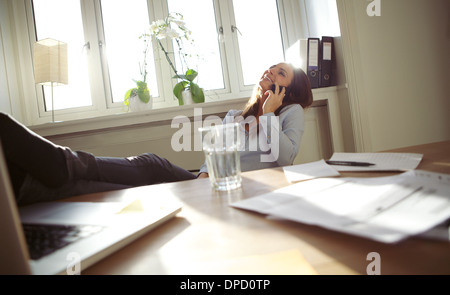 Fröhliche junge Geschäftsfrau sitzt an ihrem Schreibtisch mit Beinen auf Tisch telefonieren mit Handy. Kaukasische junge Frau entspannend Stockfoto