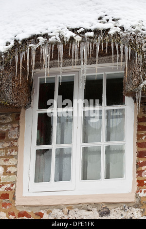 Eine Reihe von Eiszapfen hängen von einem Strohdach rund um ein Fenster auf einer Hütte Stockfoto