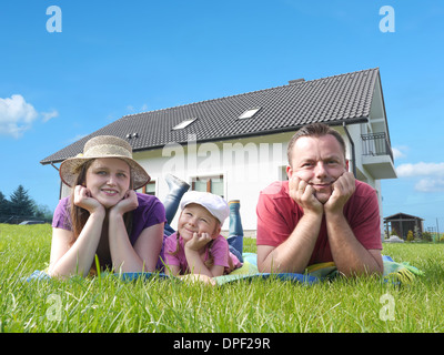 Junge Eltern mit ihren niedlichen kleinen Mädchen liegen entspannt auf dem Rasen vor dem Haus Stockfoto