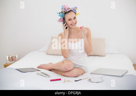 Zufrieden natürliche braune kurzhaarige Frau in Lockenwicklern Glanz anwenden, während Sie auf dem Telefon Stockfoto