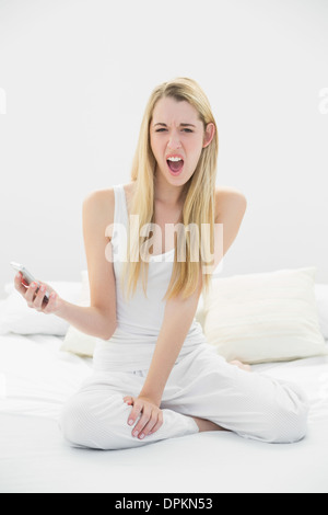 Schockiert blonde Frau, die ihr Smartphone beim Sitzen auf ihrem Bett Stockfoto