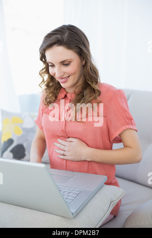 Lächelnde schwangere Frau mit ihrem Notebook auf Couch im Wohnzimmer Stockfoto