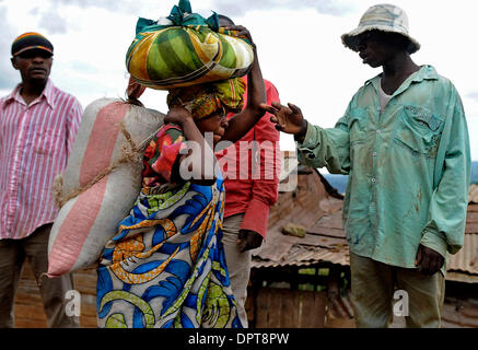 15. April 2009 - Bukavu, Kongo - A Frau tragen Bündel auf den Rücken und Kopf führt junge Männer stehen am Straßenrand in Bukavu, Kongo. In der kongolesischen Kultur Frauen scheinen, alles zu tun: die Felder zu arbeiten, das Brennholz zu sammeln, halten das Haus, die Kinder erziehen und kochen die Mahlzeiten. Die Mehrheit der ländlichen Ehemänner scheinen relativ wenig zu tun. Allerdings ohne Männer Unterstützung und re Stockfoto