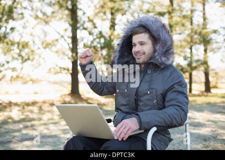 Fröhlicher Mensch im Fell Kapuze Jacke mit Laptop im Wald Stockfoto