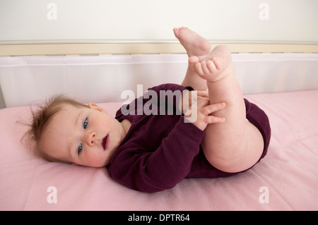 Babymädchen in ihrem Kinderbett liegen und spielen mit ihren Füßen Stockfoto