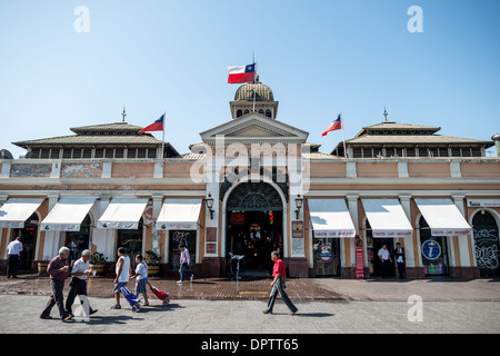 Eingang zum Mercado Central Santiago Chile | SANTIAGO, Chile — Fußgänger gehen an dem kunstvoll gestalteten Eingang des Mercado Central vorbei, einem historischen Marktplatz und kulinarischen Ziel in der Nähe des Flusses Mapocho. Das 1872 gegründete Nationaldenkmal verfügt über neoklassizistische Mauerwerke, die vom chilenischen Architekten Fermín Vivaceta entworfen wurden. Die Außenfassade des Gebäudes integriert ein charakteristisches Gusseisen-Gefüge mit neoklassizistischen architektonischen Elementen, betont durch ein zentrales Giebeldach, kunstvolle Metallarbeiten am Torbogen und eine gewölbte Kuppel. Stockfoto