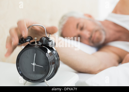 Reifer Mann streckte Hand mit Wecker im Bett Stockfoto