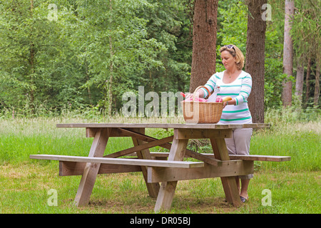 Eine Frau, die Platzierung eines Whicker Picknickkorb voller Essen und trinken auf einem Tisch im Wald, an einem hellen Sommertag. Stockfoto