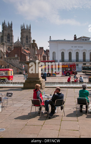 Menschen sitzen & Genießen im Freien Snack im sonnigen Piazza von Springbrunnen und Statue, Türme von York Minster & De Grau Zimmer darüber hinaus - York, England, UK. Stockfoto