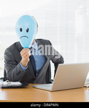 Geschäftsmann mit traurigen Smiley konfrontiert Ballon am Schreibtisch Stockfoto