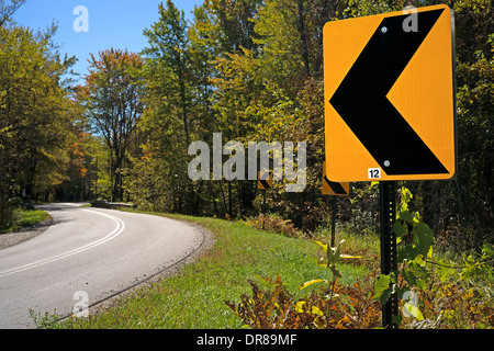 Eine deutliche gelbe und schwarze Straße Kurve voraus Zeichen im Vordergrund mit einer Straße Kurven durch den Wald im Hintergrund. Stockfoto