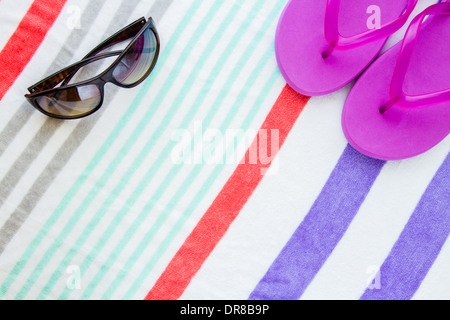 Strand-Szene mit lila Flip Flops und Sonnenbrillen auf einem gestreiften Strandtuch. Stockfoto