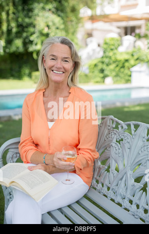 Lächelnde senior Frau Buch lesen und Weintrinken im Hinterhof Stockfoto