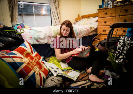 Eine junge Frau Studentin studiert, arbeitet an ihrem Laptop-Computer in ihrem chaotisch Schlafzimmer zu Hause UK Stockfoto